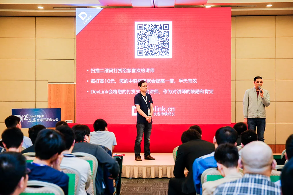 杨永全站在台上，手持话筒讲话，鸟哥站在旁边，台下坐着几百个程序员